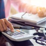 A man enters numbers on a calculator with a stethoscope lying on a desk to indicate healthcare payment integrity