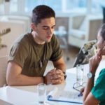 A member of the US Army sits at a table and discusses the the benefits of the PACT Act 2022 for veterans with a doctor.