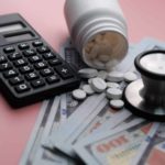 A calculator, stethoscope, and bottle of pharmaceuticals sit atop hundred dollar bills to show savings from managed care in healthcare