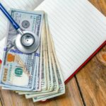 The value of cost containment in healthcare is illustrated by a stethoscope atop a pile of money and a blank journal on a wooden table.